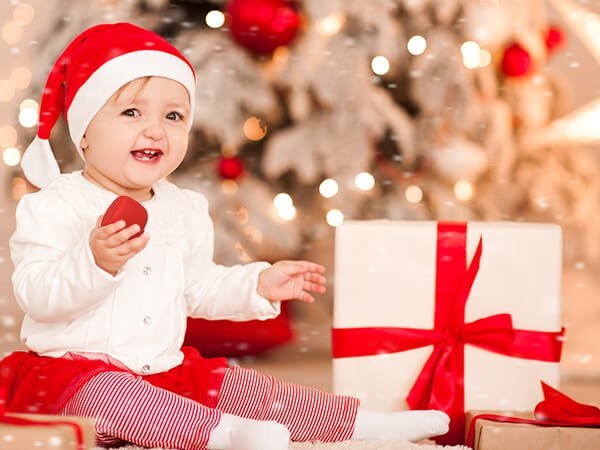 Smiling baby in Santa hat beside gifts in a festive room, Babelio holiday nursery inspiration.