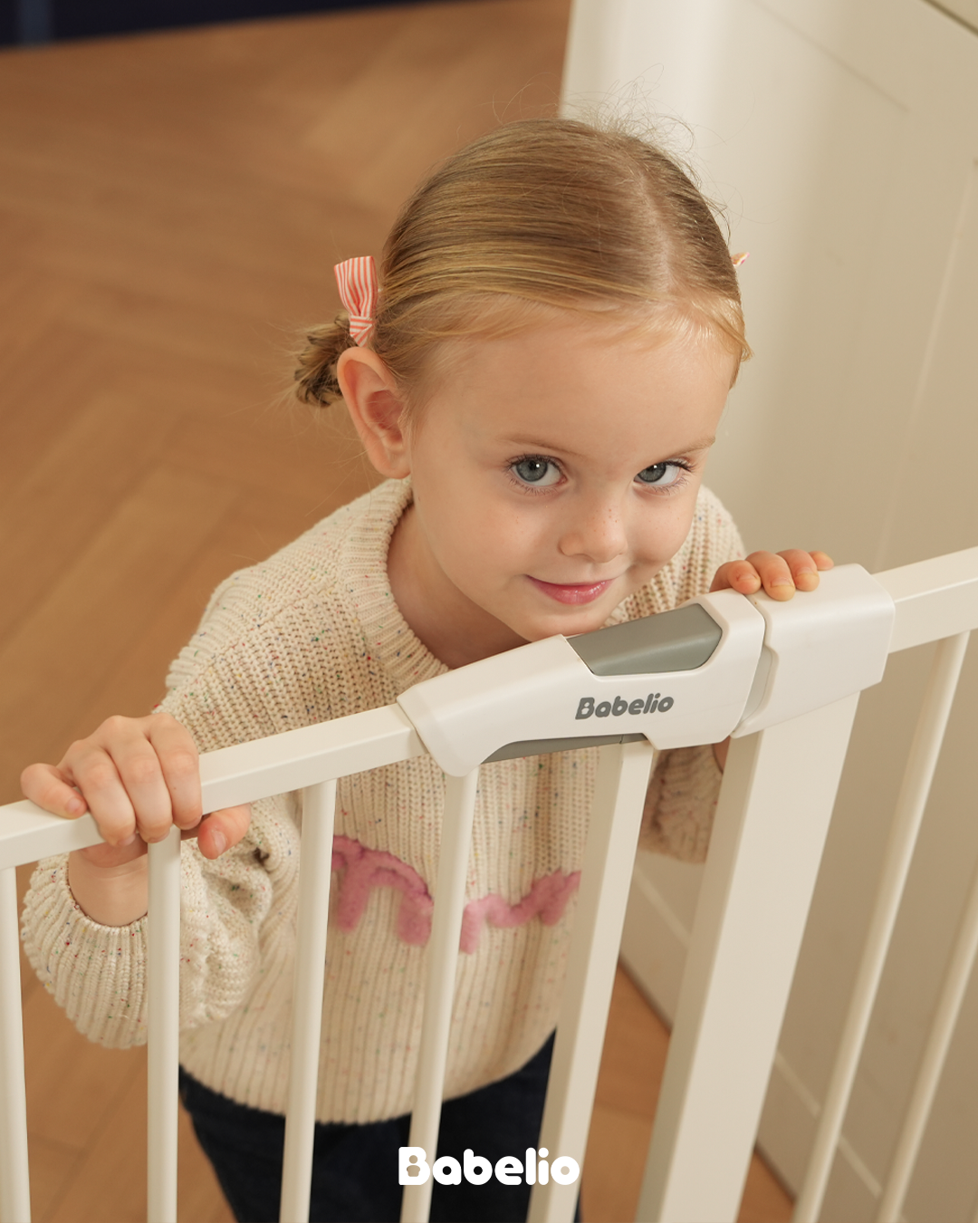 A happy toddler girl smiling in a white Babeliobaby baby gate, representing the complete Babeliobaby collection of safe nursery products.