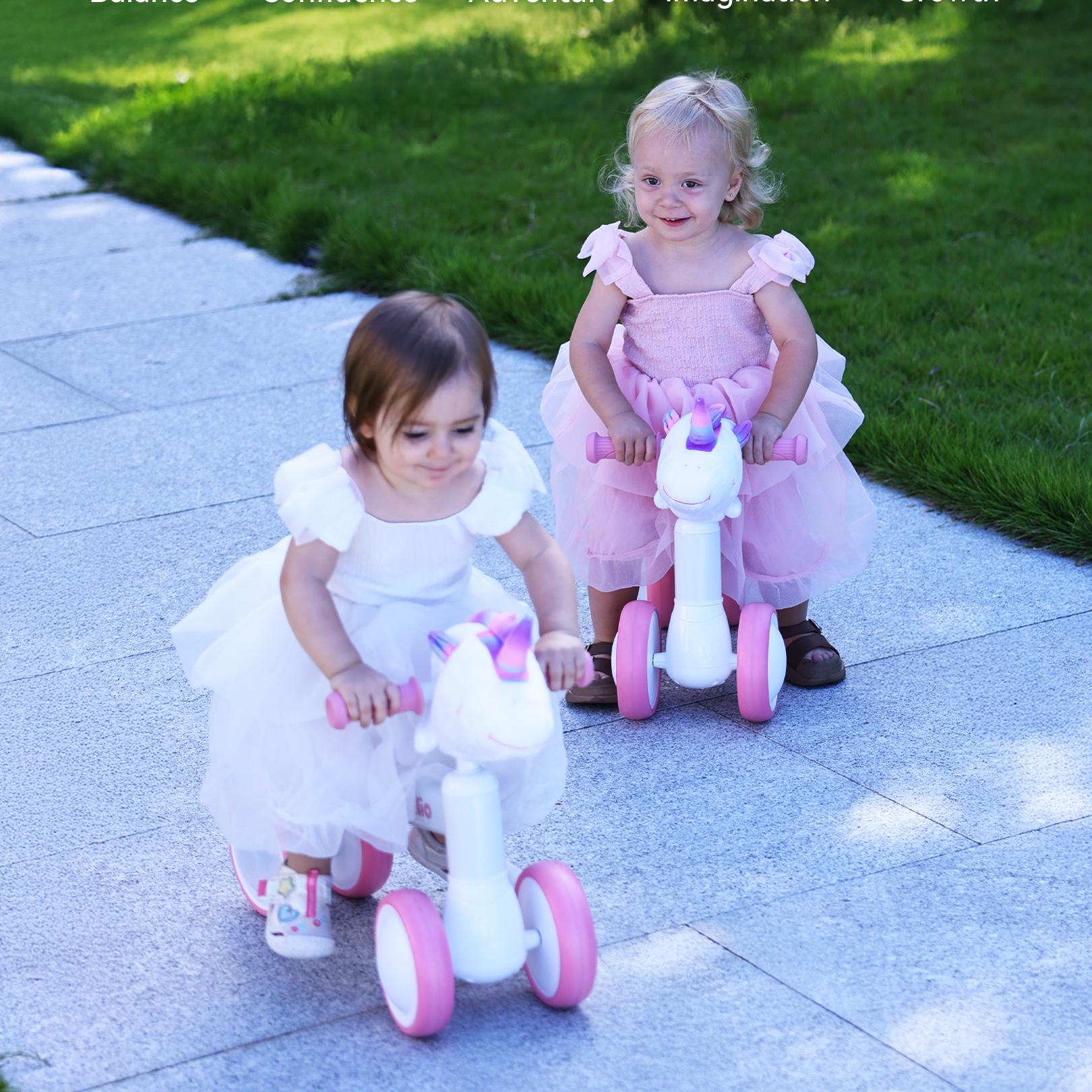Two toddlers in pink and white dresses ride pink unicorn-themed bikes outdoors, Babelio