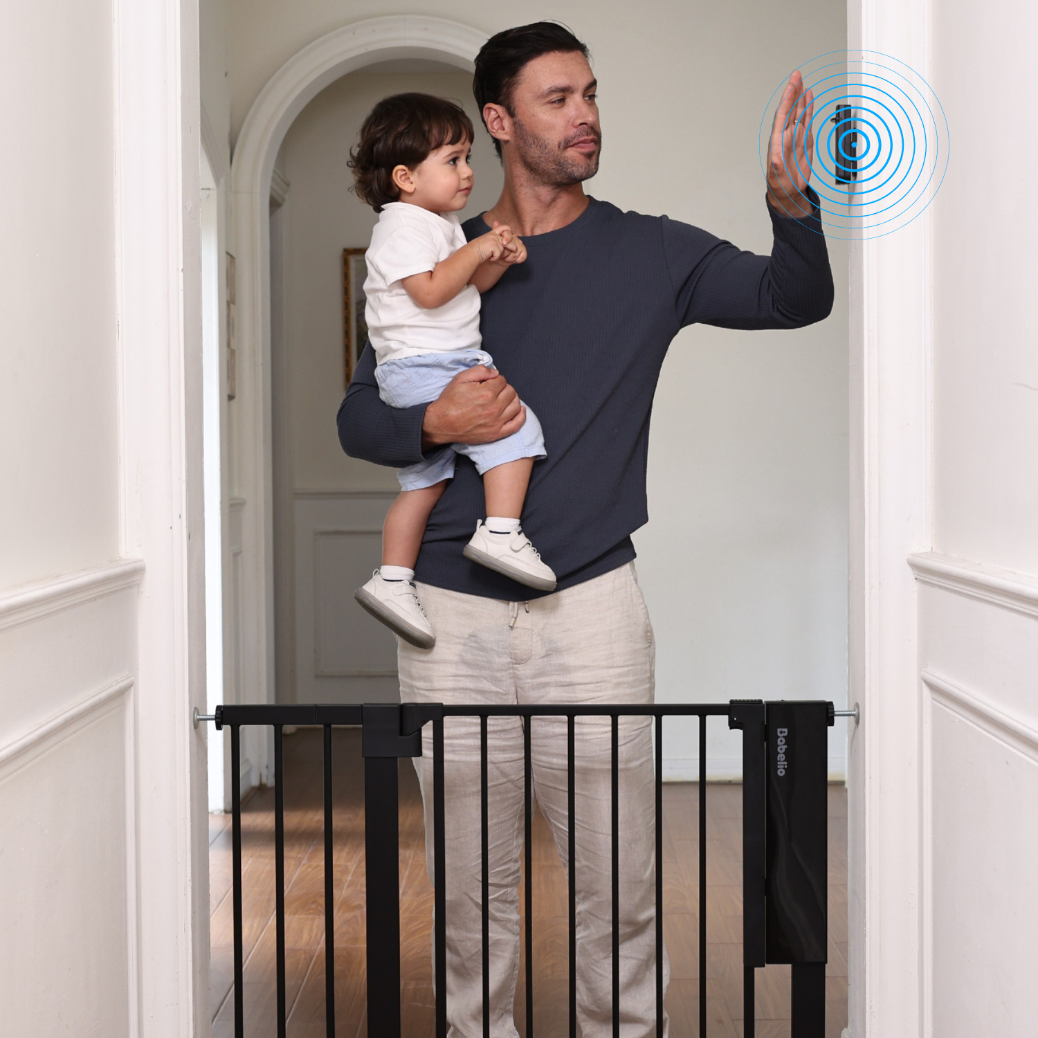 A father holding a baby, raising his hand to activate the Babelio Sensepass smart baby gate with its infrared auto-unlock feature, showing ease of use for doorways and stairs.