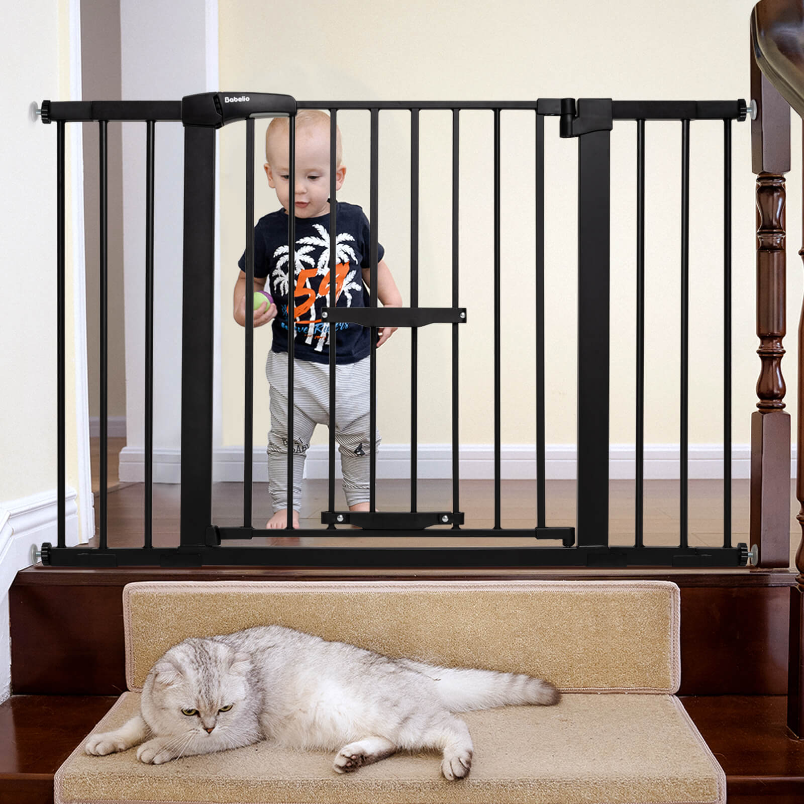 A child standing behind a black expandable baby and pet gate with a cat lying down in front of the gate on a staircase.