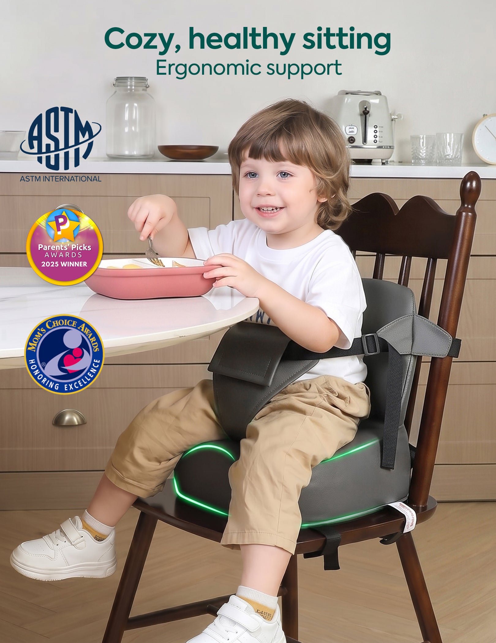 A young child sitting at a wooden dining chair wearing a dark gray ergonomic child seat belt with green LED lighting accents. The scene is set in a bright kitchen with a table and cabinets in the background, featuring promotional text and award badges.