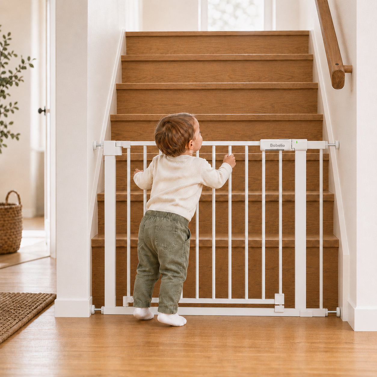 A toddler stands at a white safety gate across a wooden staircase in a bright home interior.#Color_White