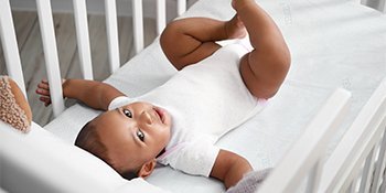 Baby in white onesie lying in crib on soft mattress, babeliobaby nursery safety products