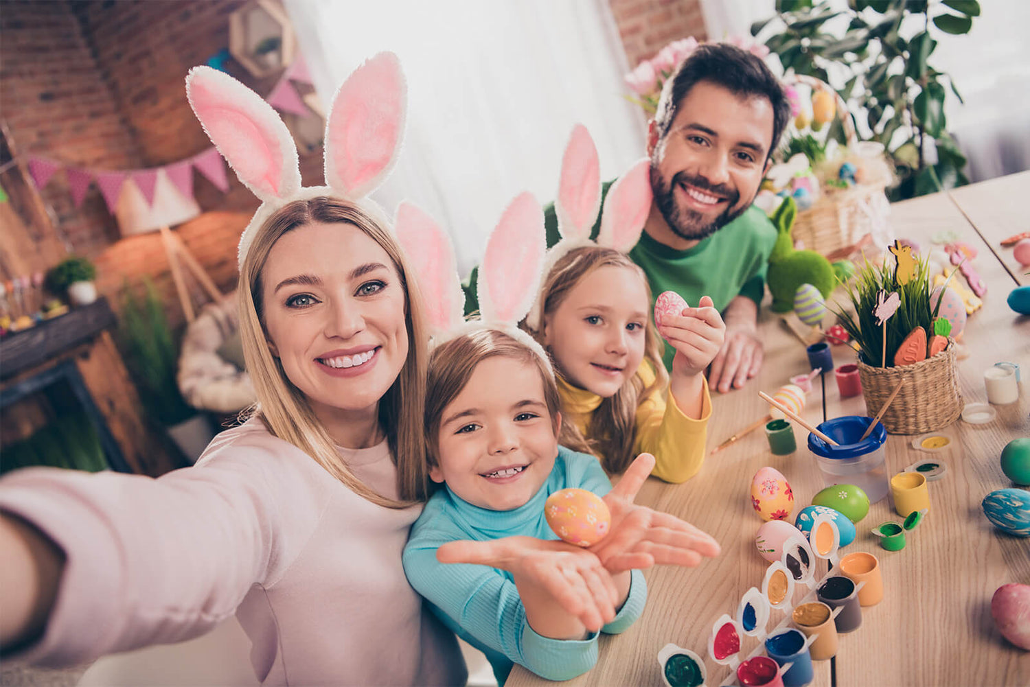Family with kids wearing bunny ears painting Easter eggs at home, surrounded by art supplies.