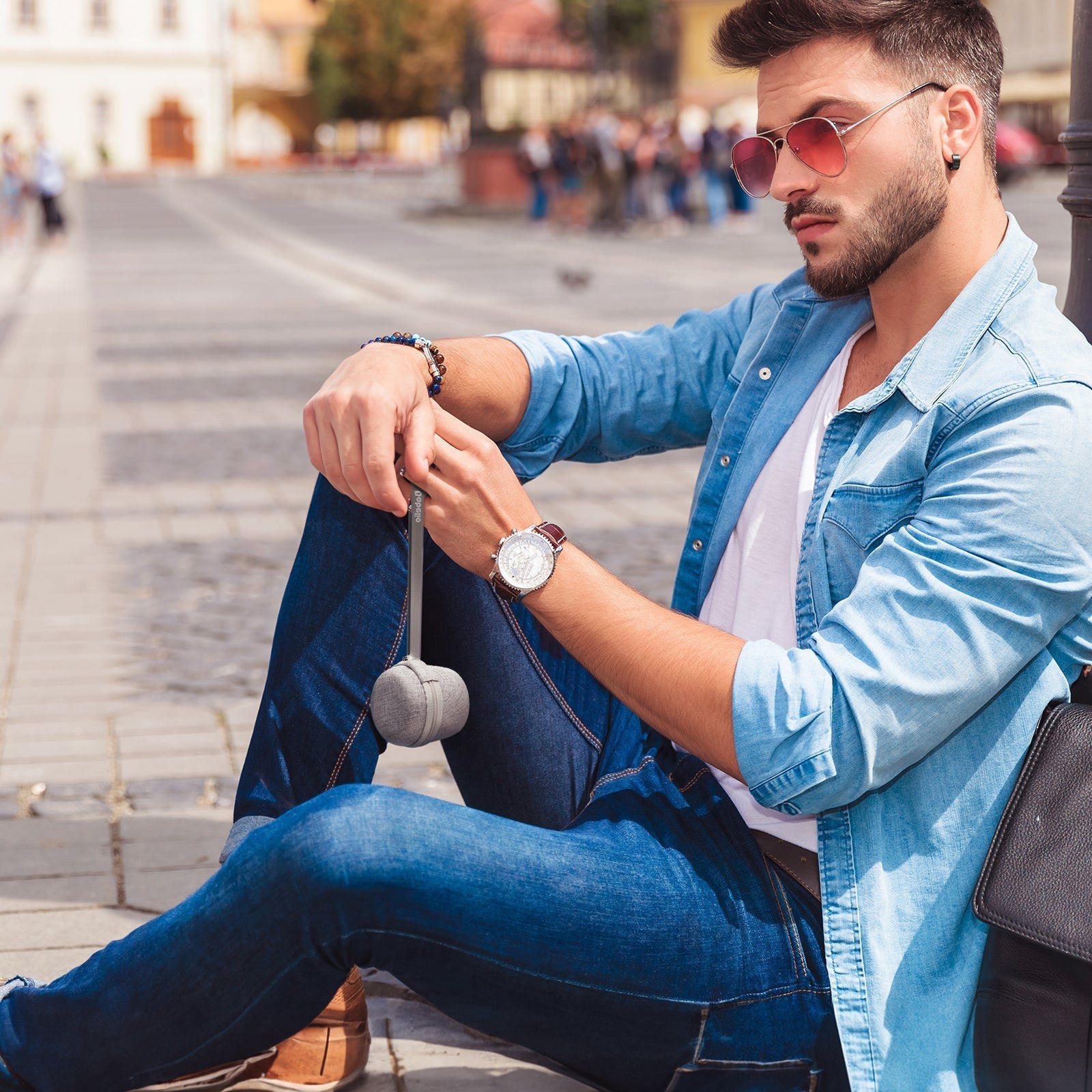Man in casual denim outfit with sunglasses and watch sitting outdoors holding a small speaker