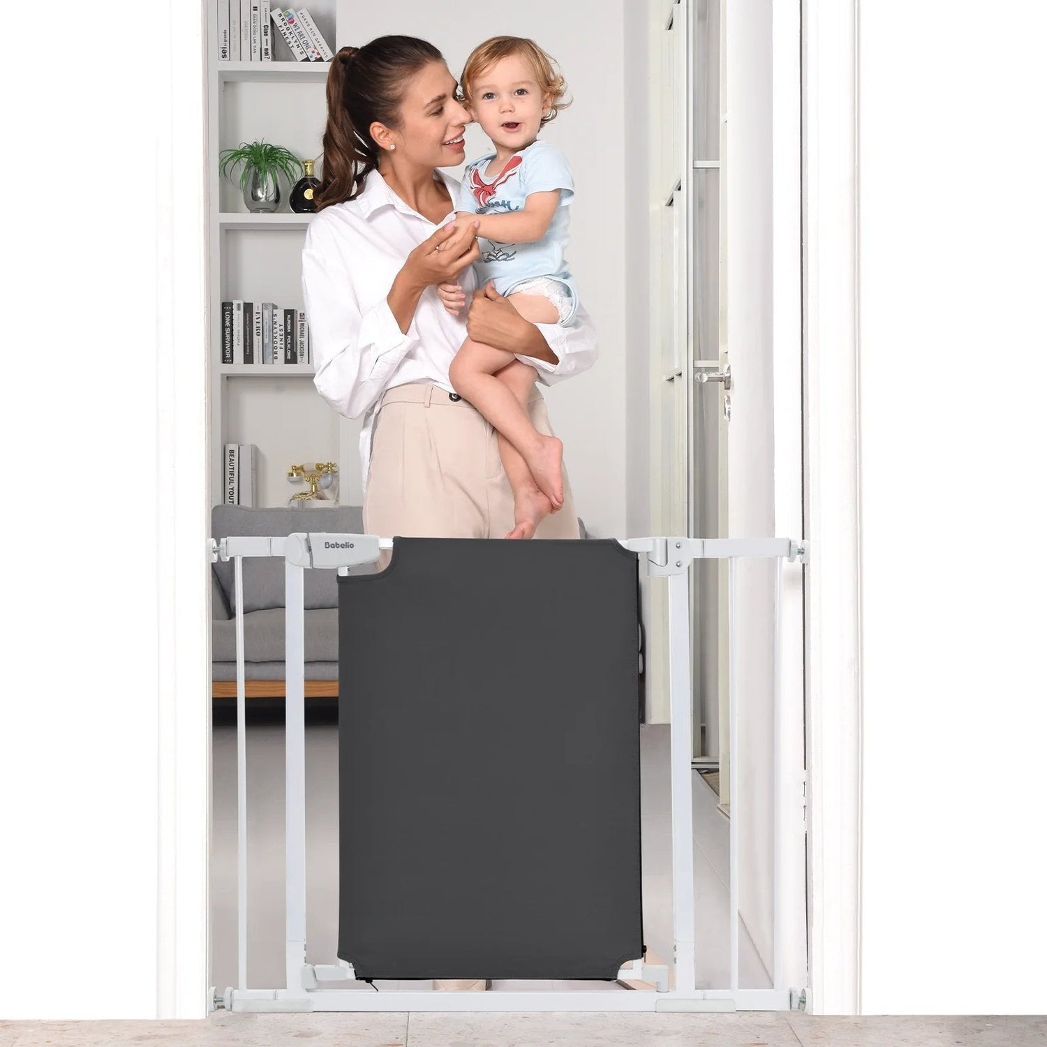 Smiling mother holds baby behind a Babelio safety gate in a modern home setting.