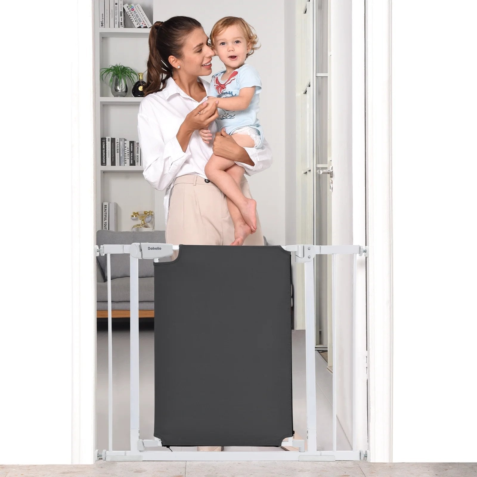 Smiling mother holds baby behind a Babelio safety gate in a modern home setting.