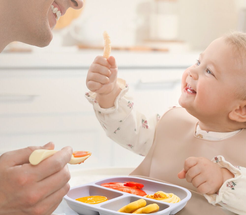Smiling baby eating from divided children’s dinnerware with parent in modern kitchen