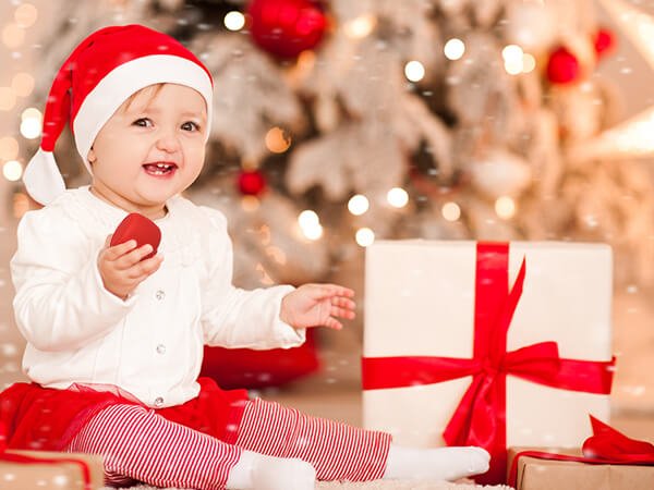 Smiling baby in Santa hat with wrapped gifts and Christmas tree, holiday scene from babeliobaby