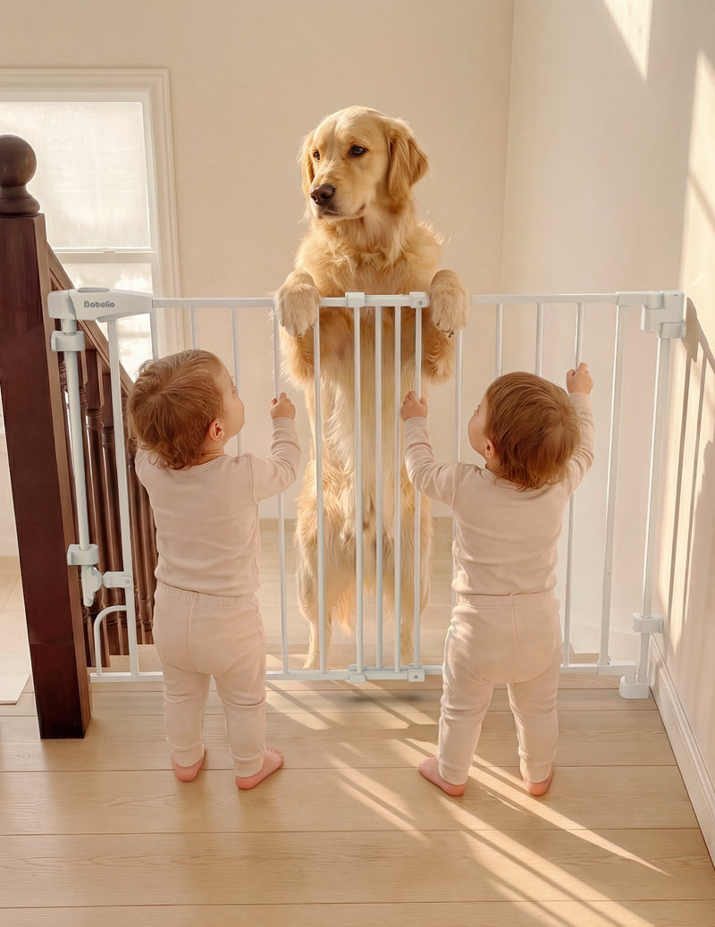 A white stair gate installed on an indoor staircase, with a golden retriever standing behind it and two toddlers in matching light pink outfits reaching up to close the gate.#Color_White