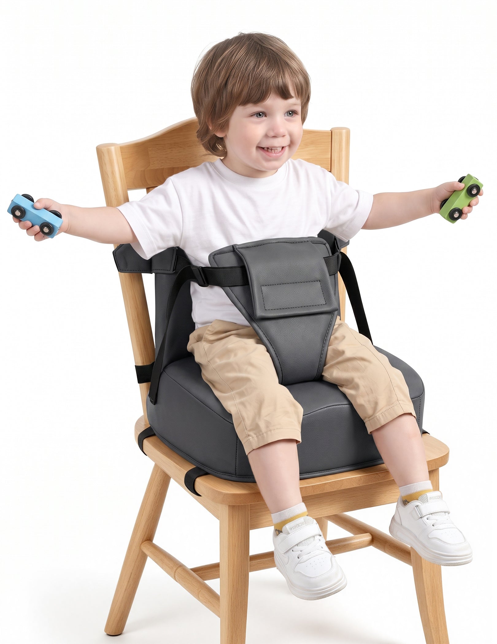 A young child sitting on a wooden chair with a dark gray padded seat belt harness attached, holding two small toy cars against a clean white background.