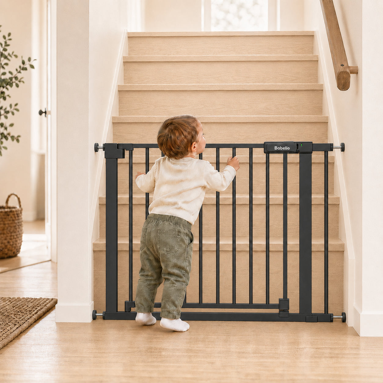 A toddler stands behind a black metal stair gate on a wooden staircase inside a bright home. The gate has a small white Brinida logo plate on the top rail.#Color_Black