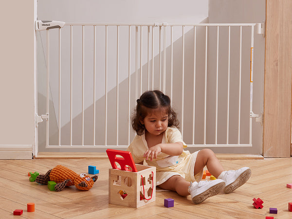 A toddler playing with toys in front of an adjustable baby gate for indoor safety, featuring auto close, no bottom bar, and sturdy drill-mounted design, Babelio Boundless B17