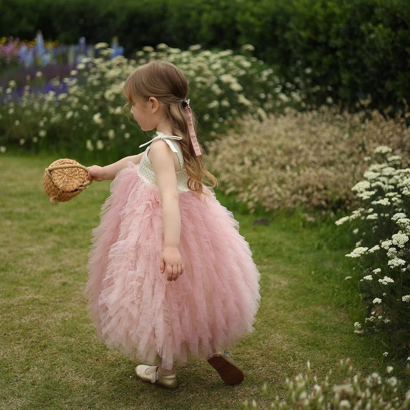 Young girl in pink tulle dress holds basket in a lush garden, flowers blooming around